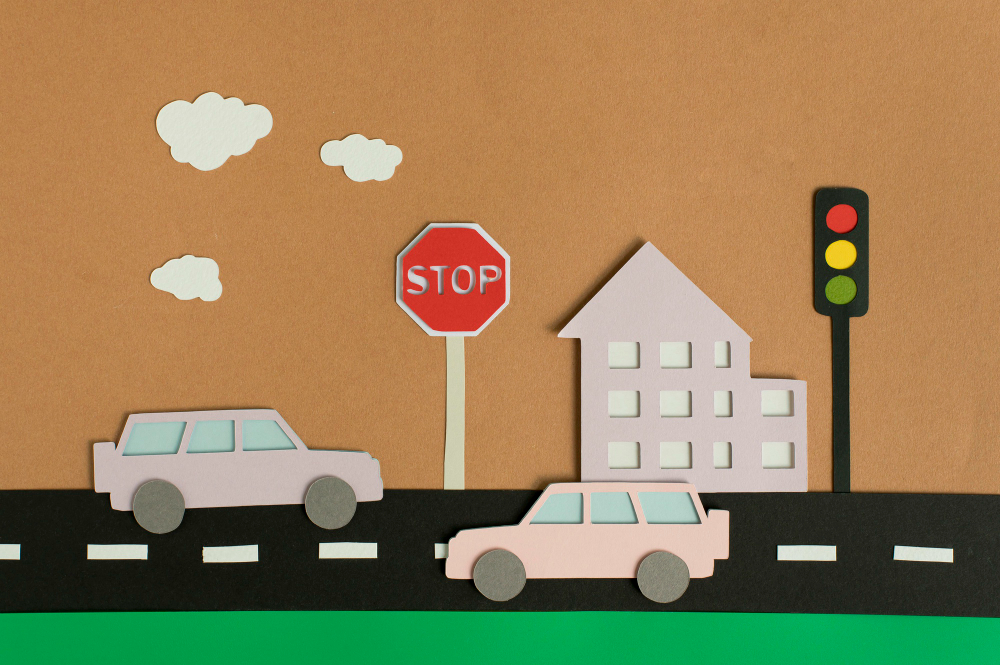 A paper art scene of a traffic calming example. There is a street with two cars, a stop sign, and a traffic light. The street is lined with a building in the background, while the sky has cut-out clouds. The traffic light shows red, yellow, and green signals, and the stop sign is prominently placed near the road.